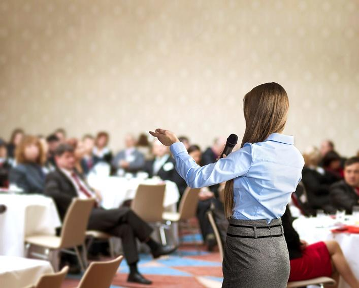 Speaker addresses an audience at a conference or business event, holding a microphone and gesturing toward seated attendees.